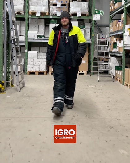 Ein lächelnder Mitarbeiter des igro-Teams läuft durch die Lagerhalle des Großmarkts. Im Vordergrund das igro Logo.