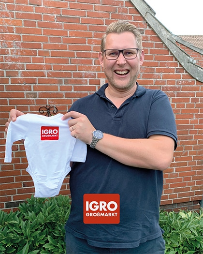 Stolzer Papa: IGRO-Mitarbeiter Dennis hält lächelnd einen weißen Baby-Body mit dem IGRO GROSSMARKT Logo in die Kamera. Im Hintergrund eine rote Backsteinwand.