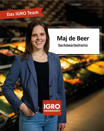 Porträtfoto von Maj de Beer im IGRO Großmarkt, engagierte Sachbearbeiterin beim igro Großmarkt. Sie trägt einen blauen Blazer, lächelt und hat die rechte Hand in der Tasche. Im Hintergrund sind unscharf Obst- und Gemüseregal zu sehen. Ein weißes Banner rechts enthält den Text „Maj de Beer, Sachbearbeiterin“. Oben links ist ein rotes Band mit „Das IGRO Team“ und unten das Logo von IGRO Großmarkt. Ein freundliches Gesicht im igro-Team.